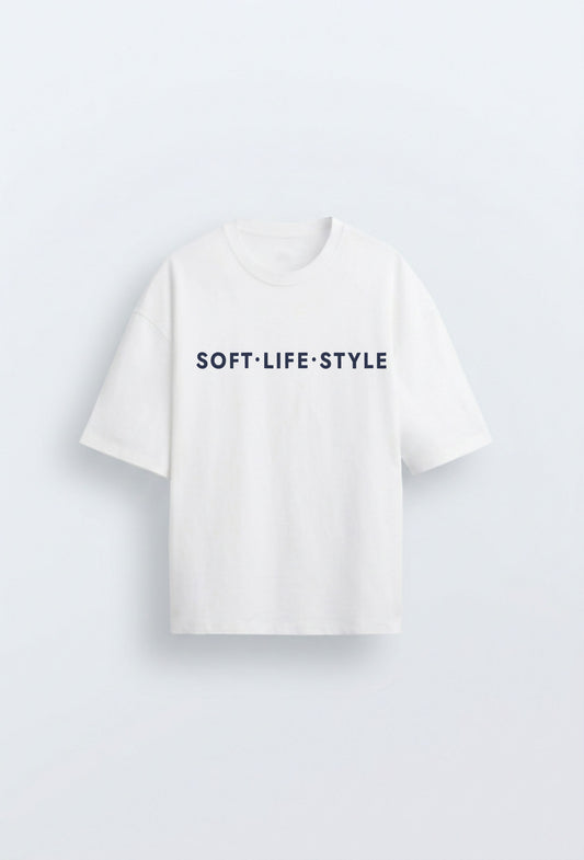 White oversized heavyweight tee with navy blue typographic print reading "SOFT·LIFE·STYLE" centred on the chest, on a clean light blue background.